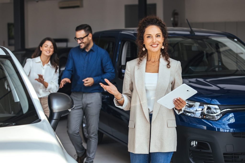 Saleswoman working at car showroom. Customers in the background. Beautiful young saleswoman working at car showroom. Professional female salesperson at car dealership.