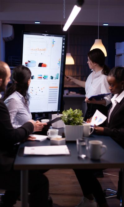 Diverse multiethnic business teamwork overworking in office meeting room analyzing financial graphs late at night. Coworkers brainstorming project ideas solving company problem.